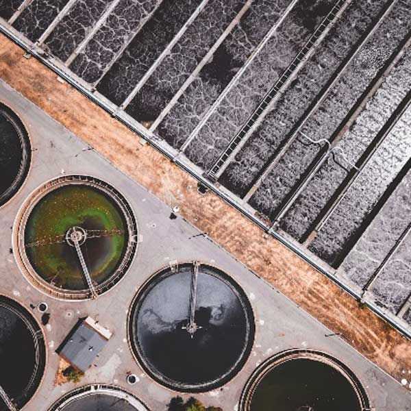 Aerial view of a wastewater treatment facility showing multiple circular settling tanks and long rectangular treatment basins separated by concrete dividers, with dark water, surface foam, and service roads visible.